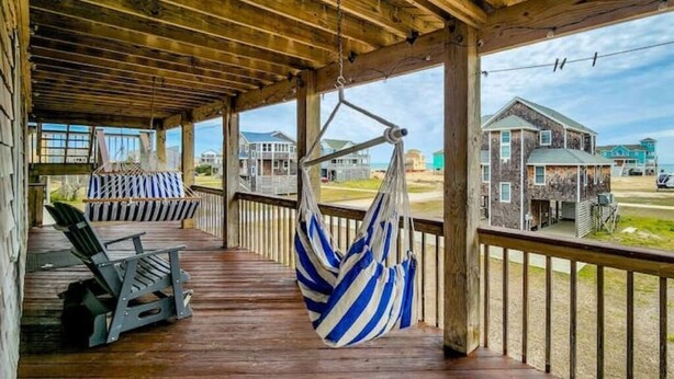 Um deck de madeira com uma rede e cadeiras, com vista para uma praia com casas.