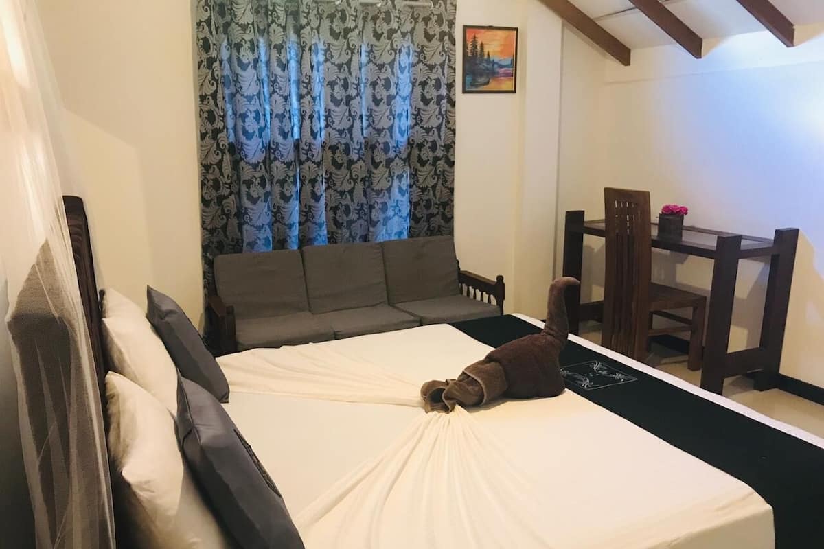Een bed met een wit laken, een bruine deken en een bruin kussen. Er staat een houten stoel en een tafel met een bloevas.