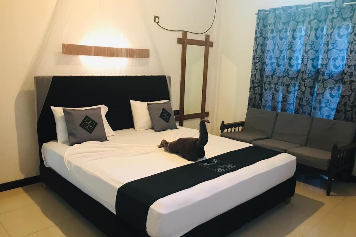 Een net opgemaakt bed met een zwart-wit deken, een houten hoofdbord, en een kat die op het bed ligt. Achtergrond toont een grijze bank en een blauw gestreept gordijn.