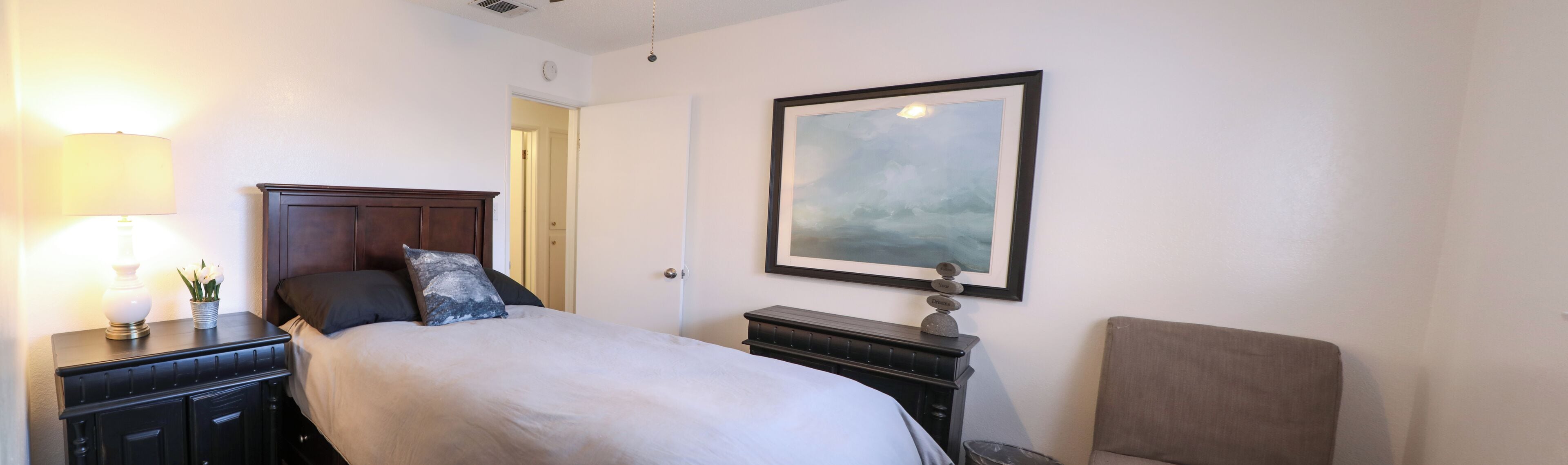 Una habitación de hotel con una cama, una mesita de noche con lámpara, un aparador y un cuadro en la pared.