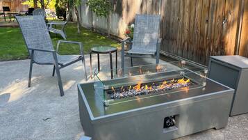 Un patio con un hogar de fuego de vidrio, dos sillas de metal, una mesita redonda y una cerca de madera al fondo.
