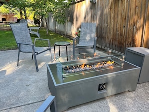 Een terras met een vuurkorf met glazen blad, twee metalen stoelen, een klein rond tafeltje en een houten hek op de achtergrond.