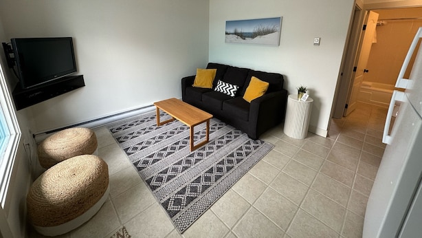 A living room with a black sofa, a wooden coffee table, two woven poufs, a patterned rug, a mounted TV, and a painting on the wall. - Boutique motel with private beach access & full kitchen (Penticton)