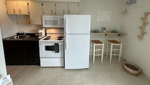 A compact kitchen with white appliances, a microwave, a stove, and a refrigerator. There are two white bar stools and a small shelf with dishes. - Boutique motel with private beach access & full kitchen (Penticton)
