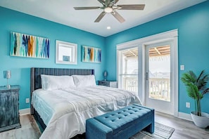 A bedroom with a bed, a ceiling fan, artwork on the walls, a potted plant, and a view through a sliding glass door.