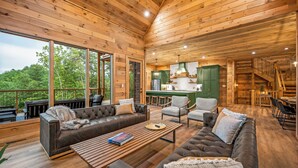 A spacious wooden cabin with a large living area featuring a sofa, armchairs, and a coffee table. A kitchen with green cabinets and a dining area with a table and chairs are visible.