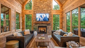 A cozy log cabin interior with a stone fireplace, leather sofas, and large windows offering a view of the forest.