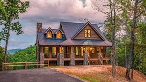 A large, two-story log cabin with a stone chimney, a wrap-around porch, and a view of the surrounding forest.