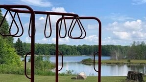 Un parque infantil con una estructura metálica roja y columpios, frente a un lago y un bosque.