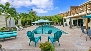 Zona de piscina con sillas y sombrillas, rodeada de palmeras y un edificio con terraza de madera.