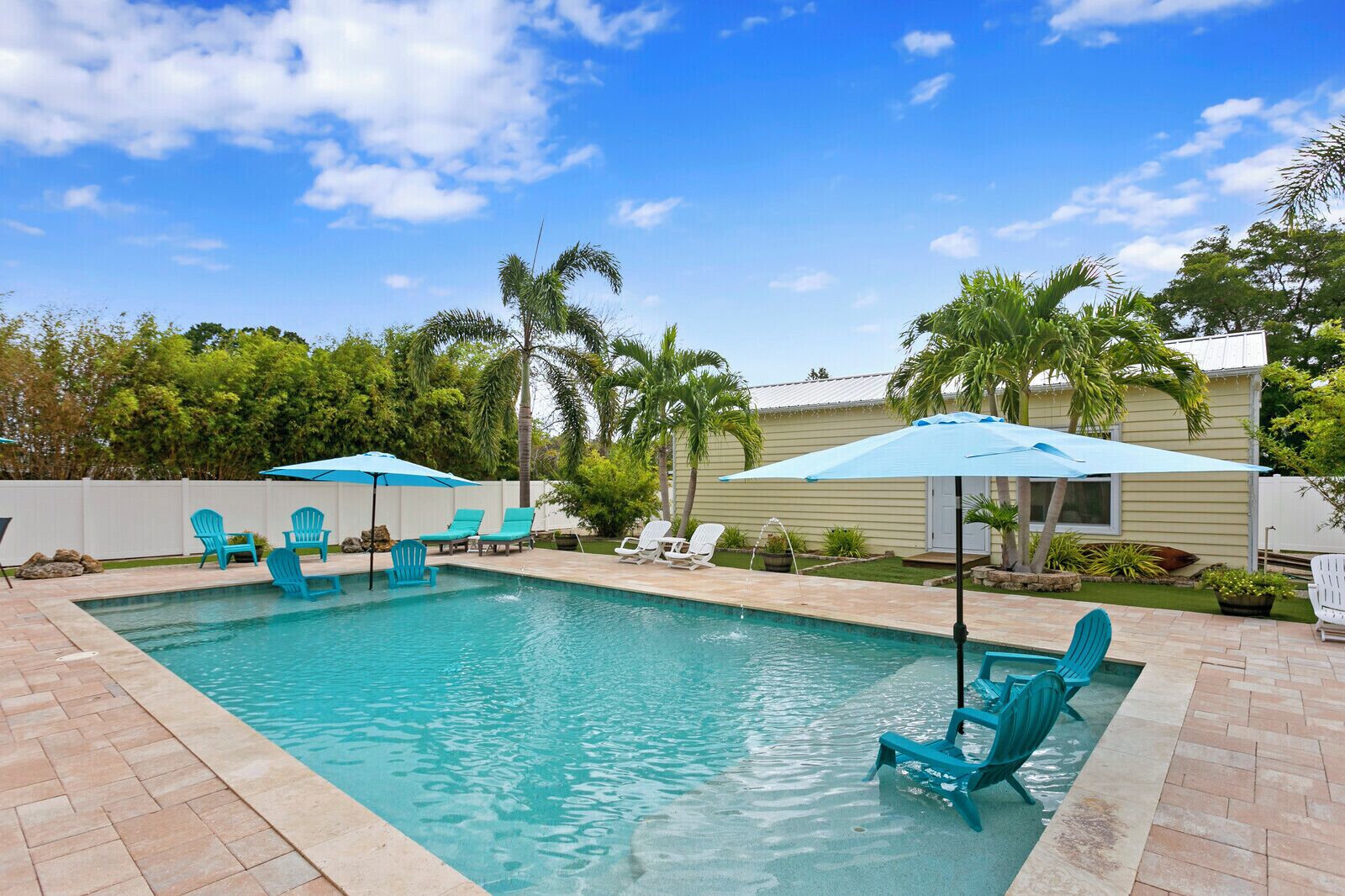 Une piscine rectangulaire à l’eau d’un bleu limpide, entourée d’une terrasse en briques. On y trouve plusieurs chaises Adirondack turquoise et un parasol bleu qui offre de l’ombre.