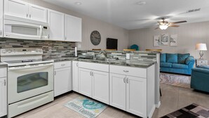 Una cocina moderna con armarios blancos, un microondas integrado y una estufa. Hay un comedor con un sofá azul y un ventilador de techo.