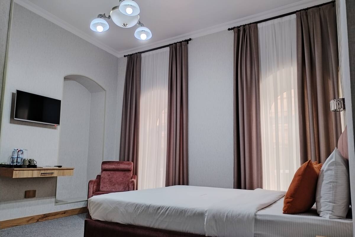 A hotel room with a bed, a desk with a flat-screen TV, a chair, and a large window with curtains.