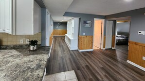 A hallway with a wooden floor, a kitchen area with white cabinets, and a bedroom visible through an open door.