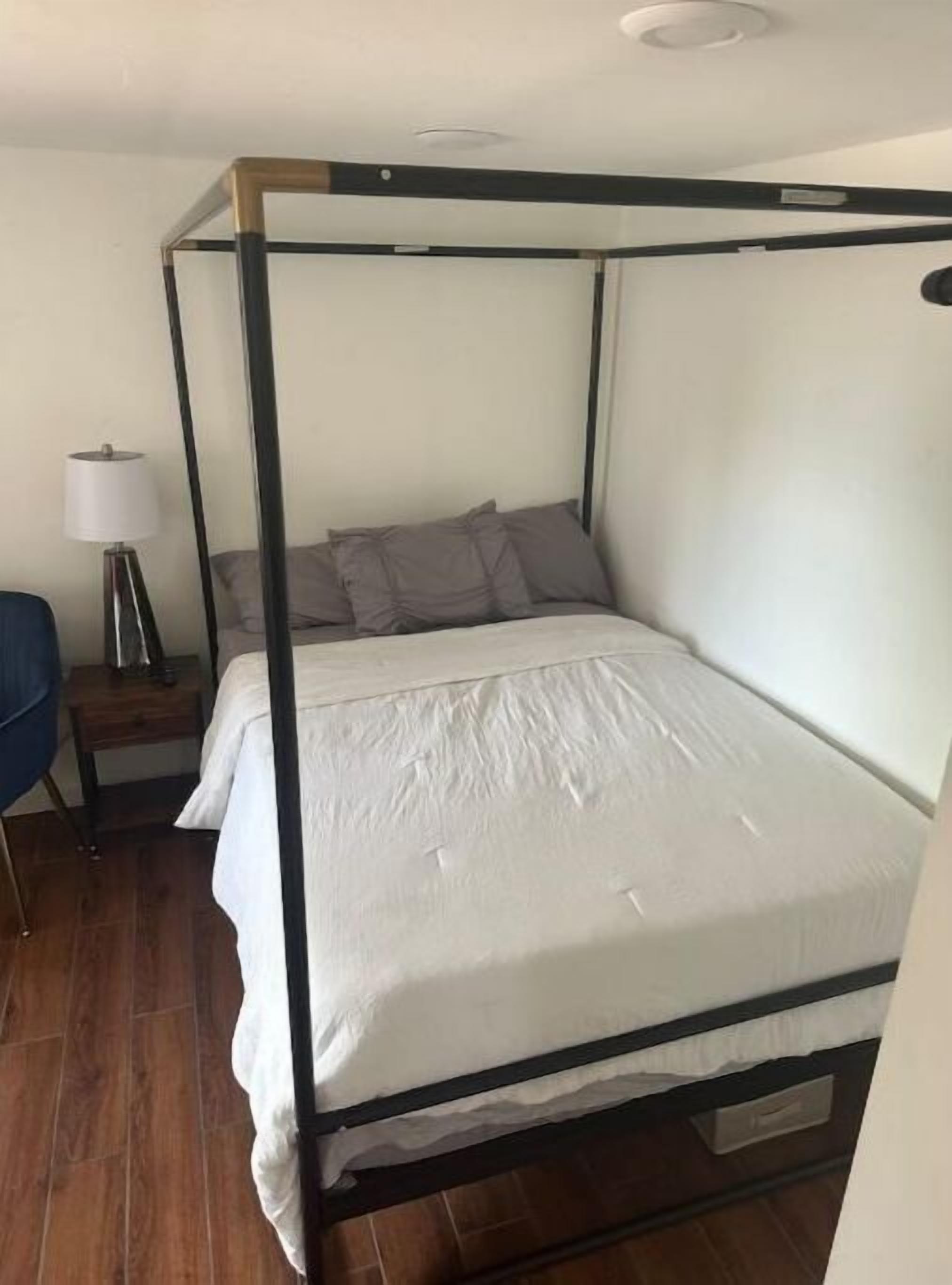 A four-poster bed with white bedding, a bedside table with a lamp, and a blue chair in a room with wooden flooring.
