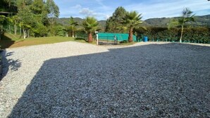 A gravel driveway leading to a fenced area with a green tarp-covered structure, surrounded by palm trees and a mountainous backdrop. - Finca-hotel Montecarlo by Hope (Antioquia)