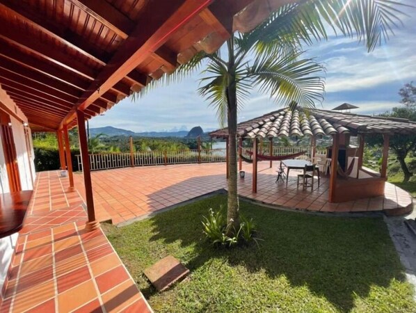 A covered patio area with a tiled floor, a wooden pergola, a seating area with tables and chairs, and a view of the surrounding landscape including hills and a palm tree.