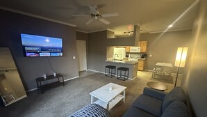 A living room with a gray sofa, a white coffee table, and a gray ottoman. There is a TV mounted on the wall, a ceiling fan, and a kitchen area in the background.