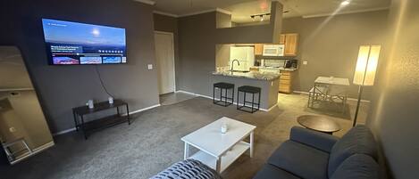 A living room with a gray sofa, a white coffee table, and a gray ottoman. There is a TV mounted on the wall, a ceiling fan, and a kitchen area in the background.