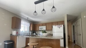 A modern kitchen with wooden cabinets, a white refrigerator, a microwave, and a sink. There are two stools at the island and a ceiling light fixture. - Peaceful Hideaway! New Listing (Roscommon)