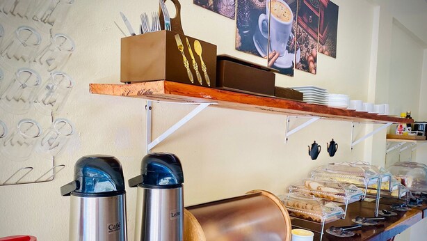 Mostrador de café con cafeteras de acero inoxidable, una exposición de pan y una obra de arte con temática de café en la pared.