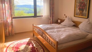 A bedroom with two beds, a large window with a view of green hills, a red patterned rug, and a framed painting on the wall.