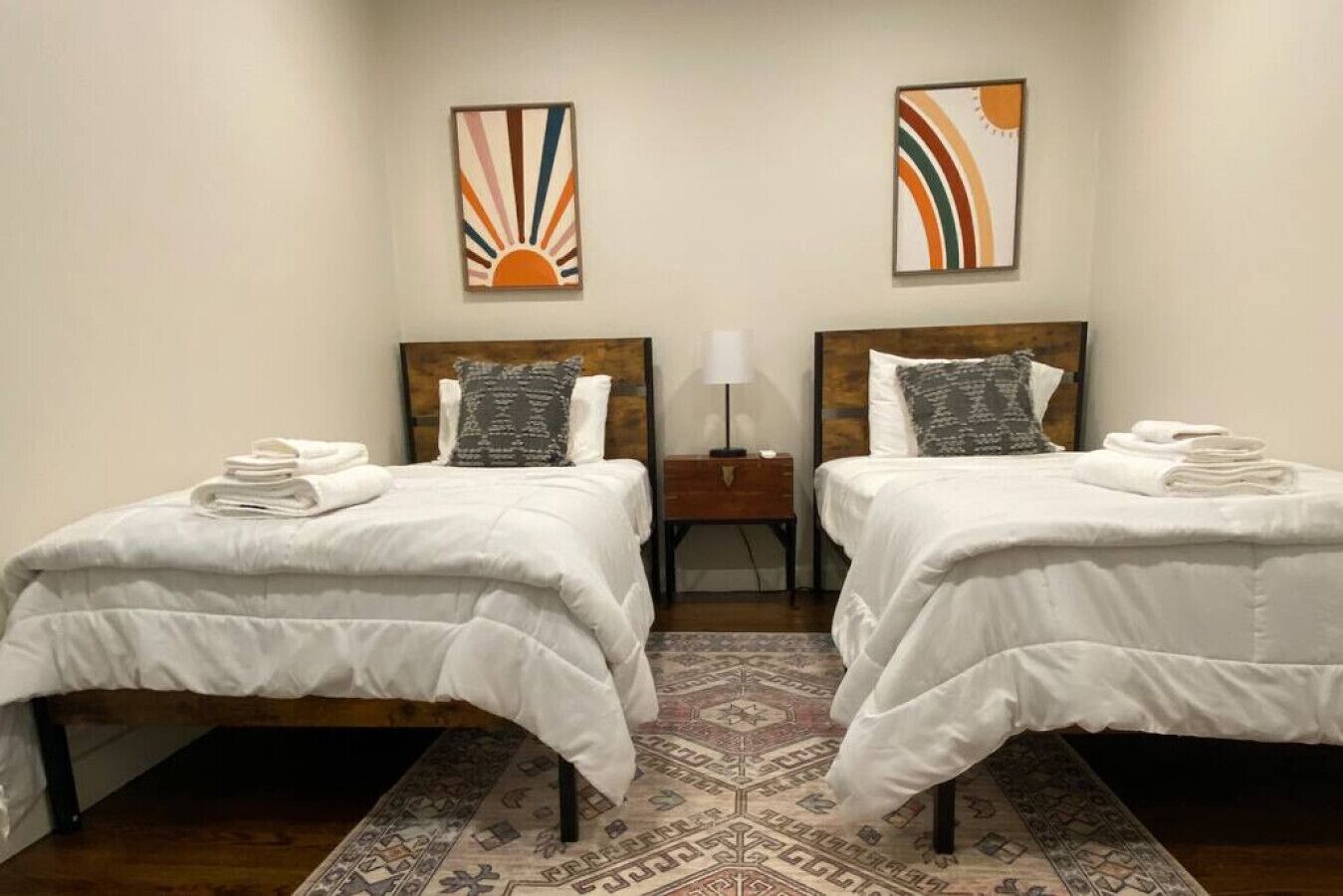 A hotel room with two single beds, each with a white duvet and a folded towel on the side. There is a nightstand with a lamp between the beds. The room has a patterned rug on the floor and two abstract paintings on the walls.