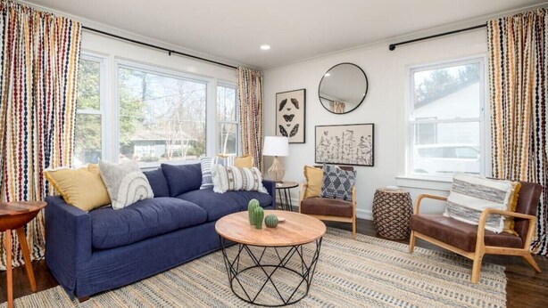 A living room with a blue sofa, a round coffee table, and a brown leather chair. There are two windows with curtains, a round mirror, and framed artwork on the wall. - Collegeside Cottage / Downtown Franklin Park/Brevard College 3Bd Pets ok (Brevard)