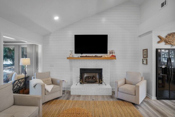 A living room with a fireplace, a flat-screen TV mounted above it, and a modern kitchen with a refrigerator. - Renovated Center Island Home & Garage Apt 4bd/3ba (Fripp Island)
