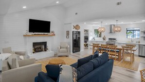 A spacious living room with a fireplace, a large flat-screen TV mounted above it, and a dining area with wooden chairs and a table. - Renovated Center Island Home & Garage Apt 4bd/3ba (Fripp Island)