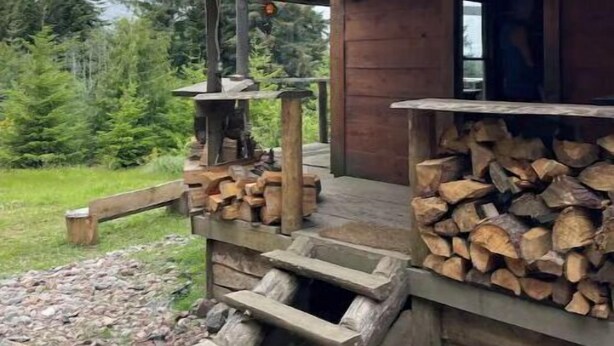A rustic cabin with a wooden porch, a fire pit, and a stack of firewood.