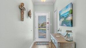 Un pasillo con piso de madera, una puerta blanca que da a vistas de edificios, un cuadro en la pared y un mueble de madera rústico con un tarro encima.