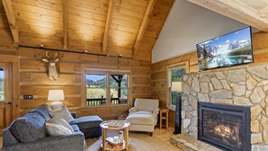 A log cabin interior with a stone fireplace, wooden ceiling beams, a mounted deer head, a television, and a view of the outdoors through the windows. - Delightful Cabin near Blue Ridge Mountains (Blowing Rock)