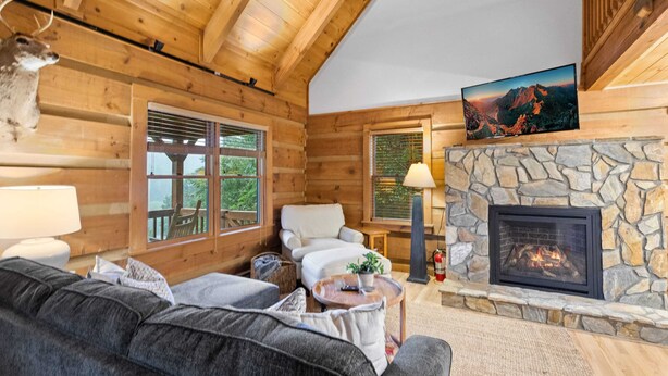 A log cabin interior with a stone fireplace, a mounted deer head, a sofa, and a television displaying a mountain scene. - Delightful Cabin near Blue Ridge Mountains (Blowing Rock)