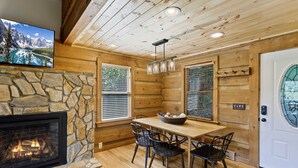 A cozy cabin interior with a stone fireplace, wooden walls, a dining table with chairs, and a large window with blinds. - Delightful Cabin near Blue Ridge Mountains (Blowing Rock)