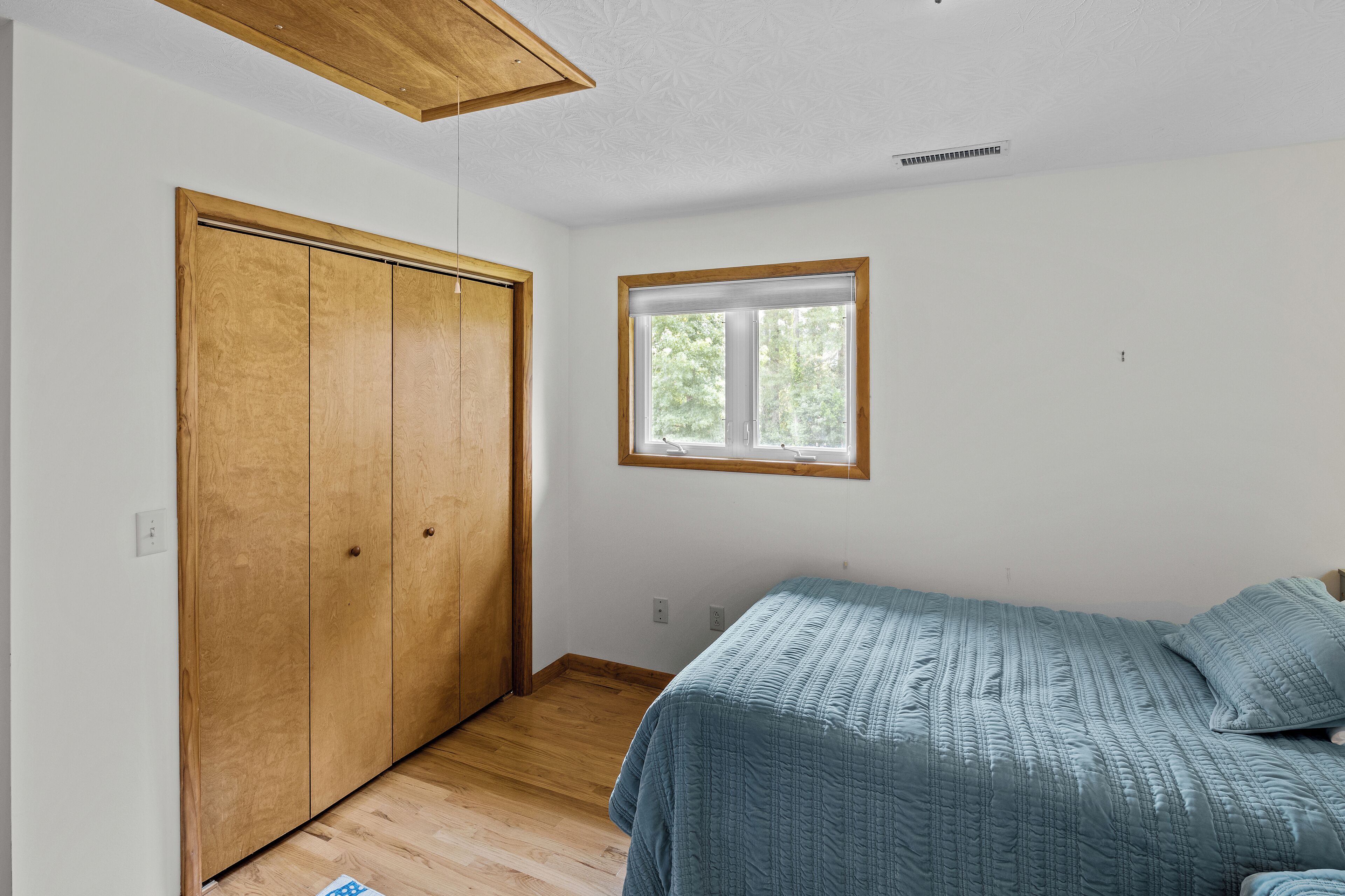Ein Schlafzimmer mit einem Holfischschrank, einem Fenster mit Blick auf Bäume und einem Bett mit blauer Bettdecke.