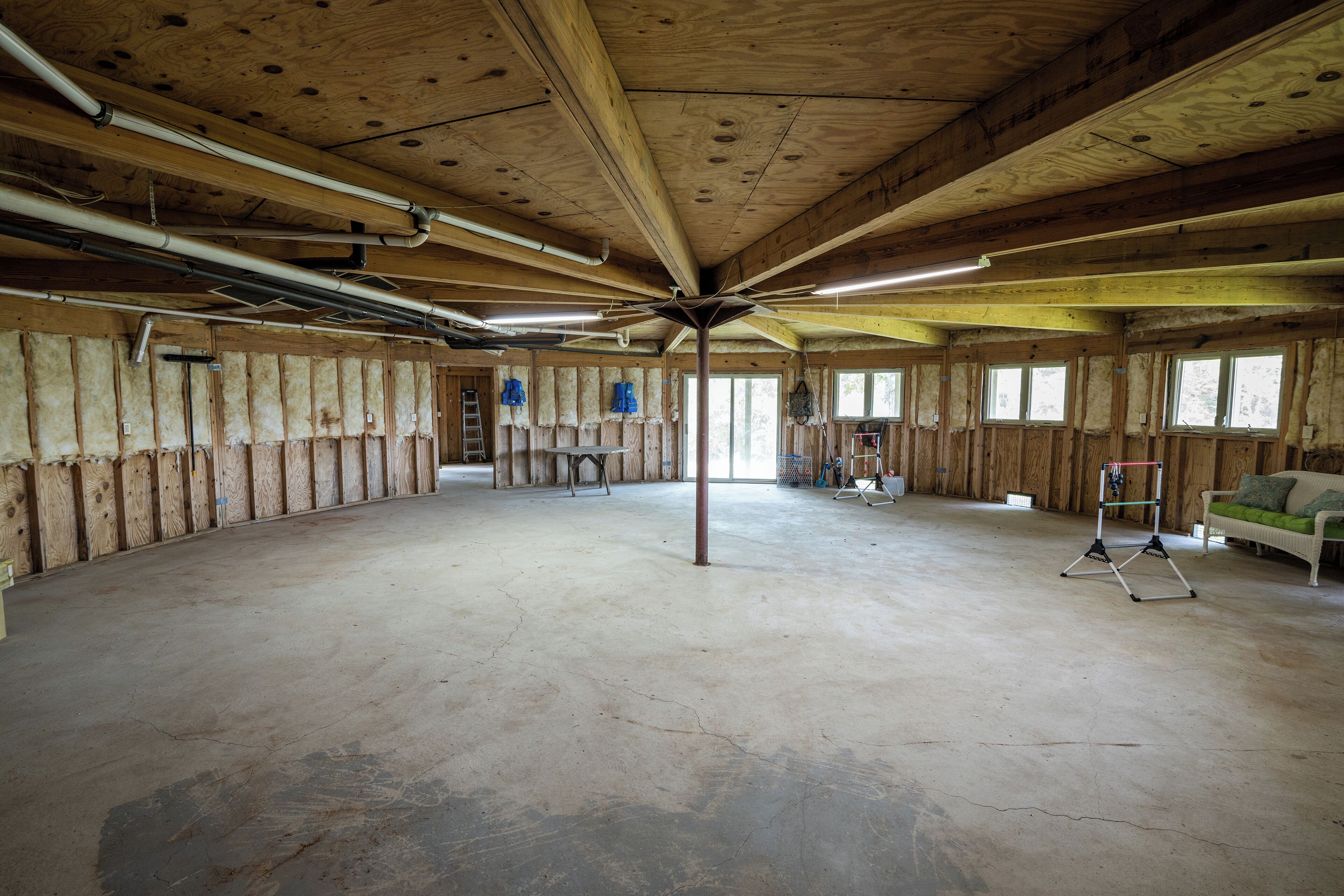 Een ruime, open kamer met zichtbare houten balken en een betonnen vloer, met een centrale kolom, een hangstoel en een kleine tafel.