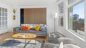 Una sala de estar moderna con un sofá gris, cojines coloridos, una mesa de centro de madera y vistas a la ciudad a través de amplios ventanales.