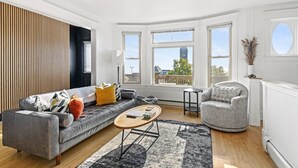 Una sala moderna con un sofá gris, una mesita redonda y un sillón gris. Amplias ventanas proporcionan luz natural y vistas al exterior.