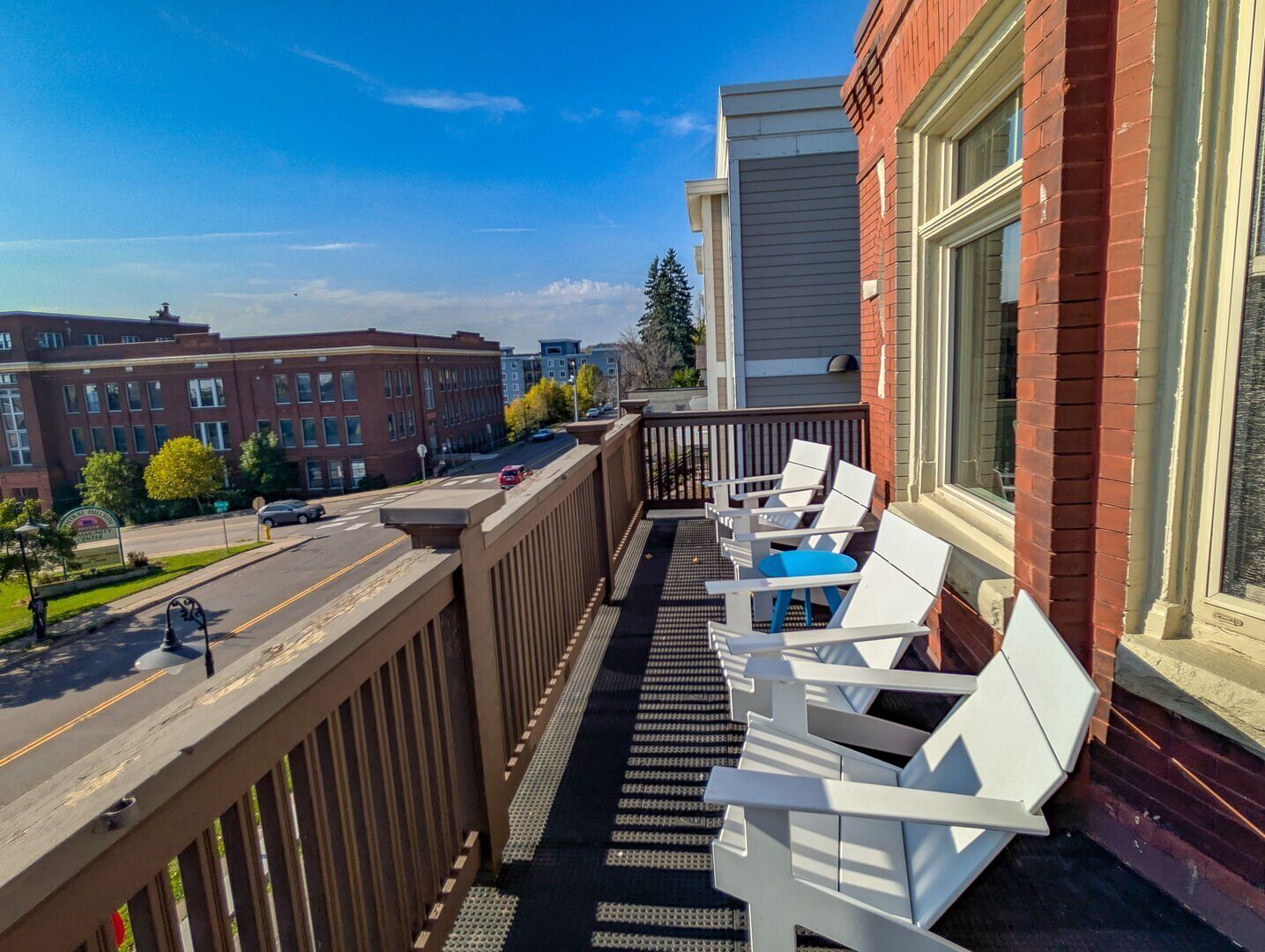 A balcony with white chairs and a blue cushion, overlooking a street with buildings and cars.