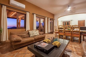 A cozy living room with a brown leather sofa, a coffee table with a suitcase and a magazine, and a dining area with wooden chairs and a table.