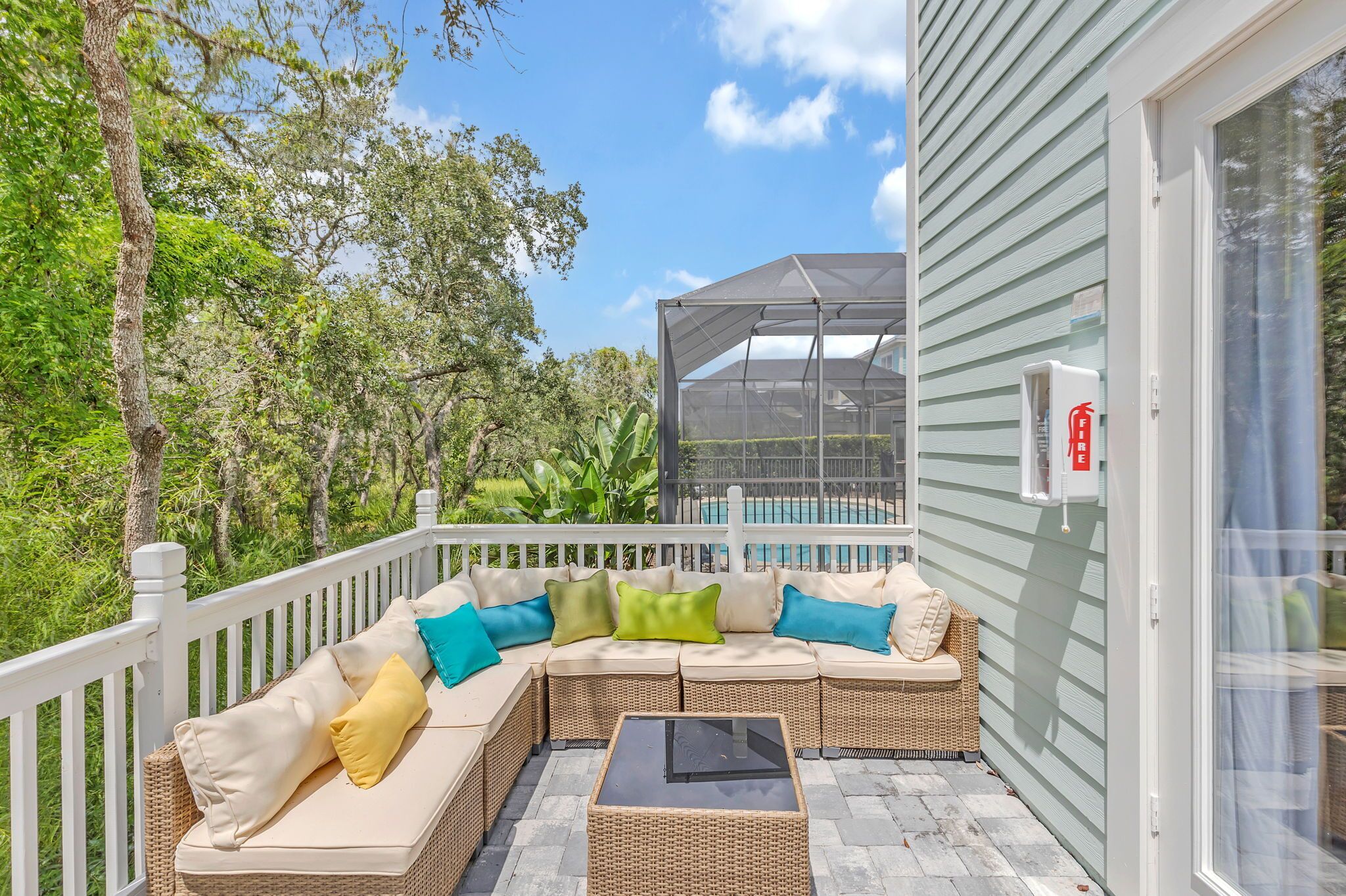 A covered outdoor seating area with a wicker sofa and cushions, a glass table, and a view of the surrounding greenery and pool.