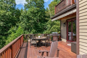 Eine Holzterrasse mit Esstisch, Stühlen, einem Grill und Blick auf dichten Wald.