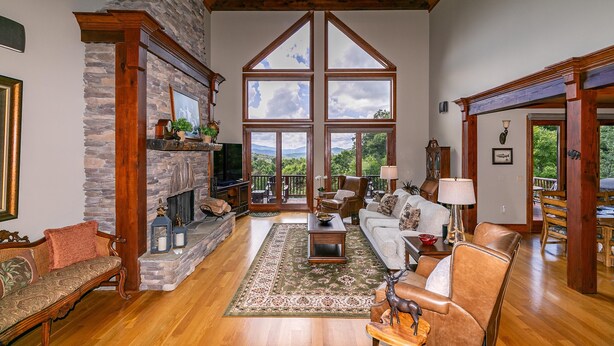 A spacious living room with a stone fireplace, wooden beams, and large windows offering a view of the outdoors. - Haven at Headwaters: Long-range Mountain Views, Pet Friendly, Gameroom (Elk Park)