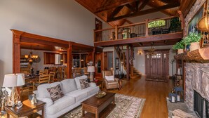 A spacious living area with a high ceiling, wooden beams, a fireplace, and a dining area with a kitchen in the background. - Haven at Headwaters: Long-range Mountain Views, Pet Friendly, Gameroom (Elk Park)