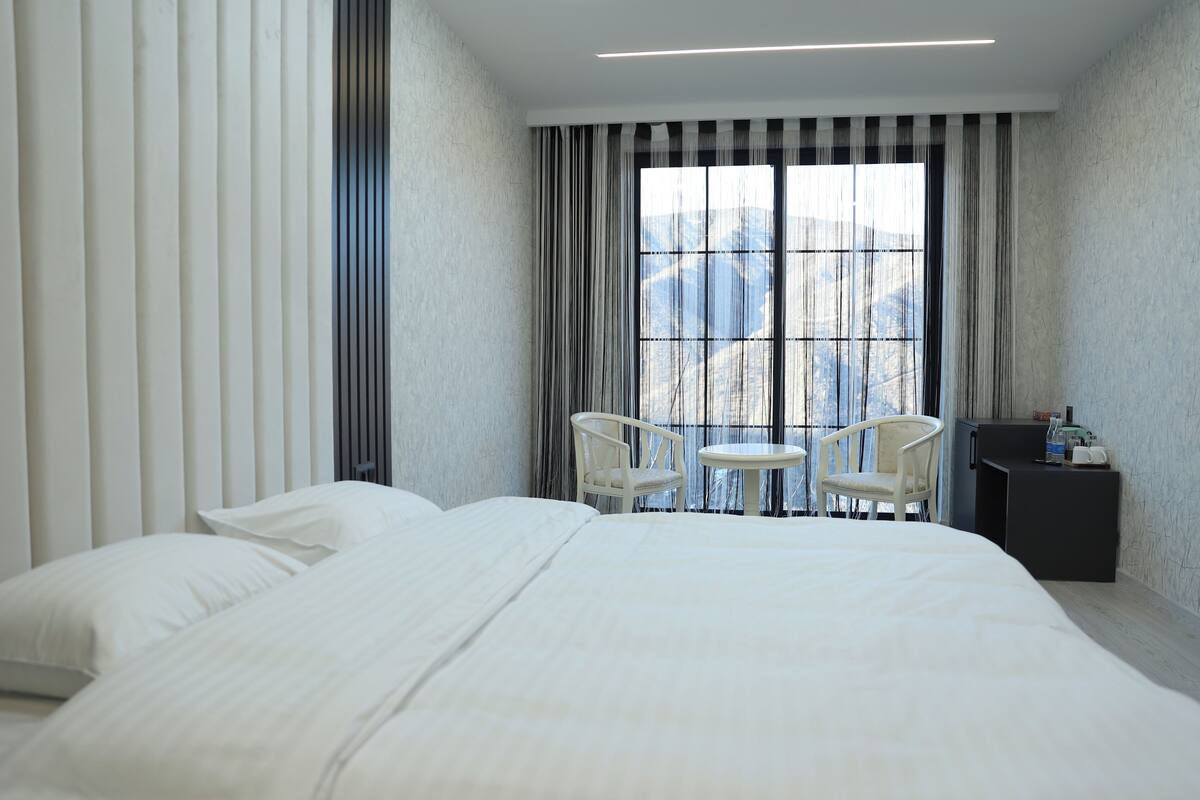 Habitación de hotel con una cama grande, dos sillas, una mesita y vistas a las montañas a través de la ventana.