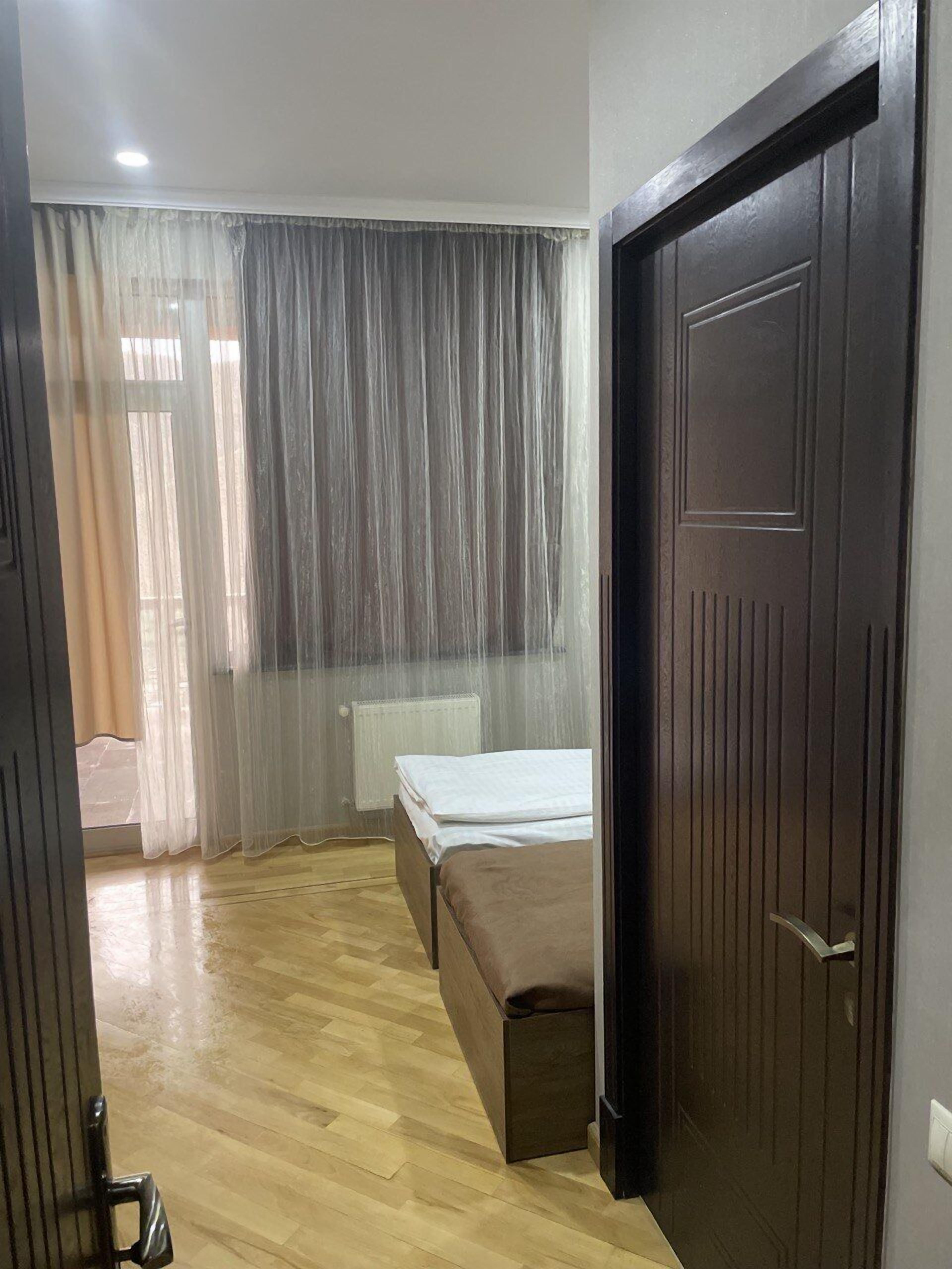 A bedroom with a wooden floor, a bed with a brown headboard, a bedside table, and a window with sheer curtains.