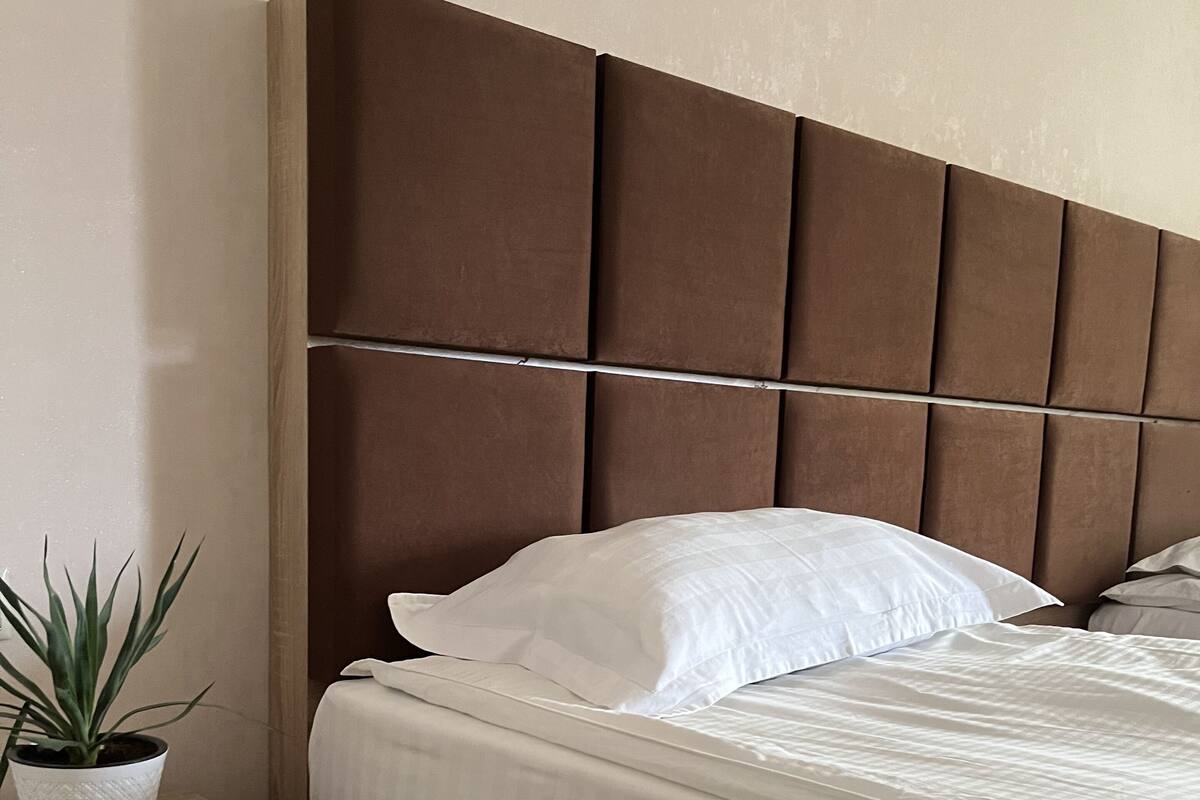 Una cama bien hecha con sábanas blancas, una mesita de noche de madera con una planta en maceta y un cabecero con diseño acolchado.