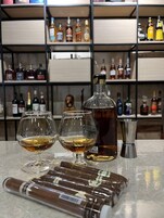 Zona de bar con dos vasos de whiskey, un decantador y un shaker metálico sobre una encimera de mármol. Al fondo, estantes repletos de diversas botellas de licor.