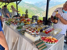 Una mesa de buffet con diversas frutas, postres y bebidas, con montañas al fondo.